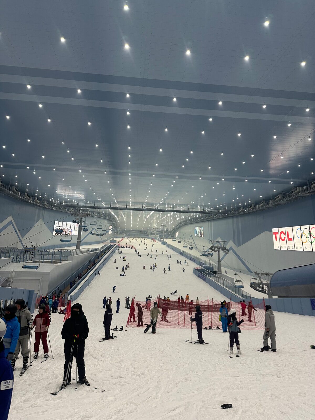 Insidan av en mycket stor inomhusskidbacke i Shenzhen. En pist med liftar sträcker sig uppåt genom hallen under ett upplyst tak, och dussintals skidåkare och snowboardåkare rör sig i backen och på planen längst ner.
