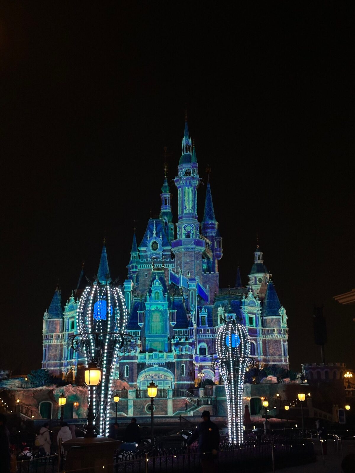 Slottet i Shanghai Disneyland upplyst i blått och lila mot en mörk natthimmel, med dekorerade lyktstolpar med vita ljusslingor i förgrunden och siluetter av besökare vid slottets fot.