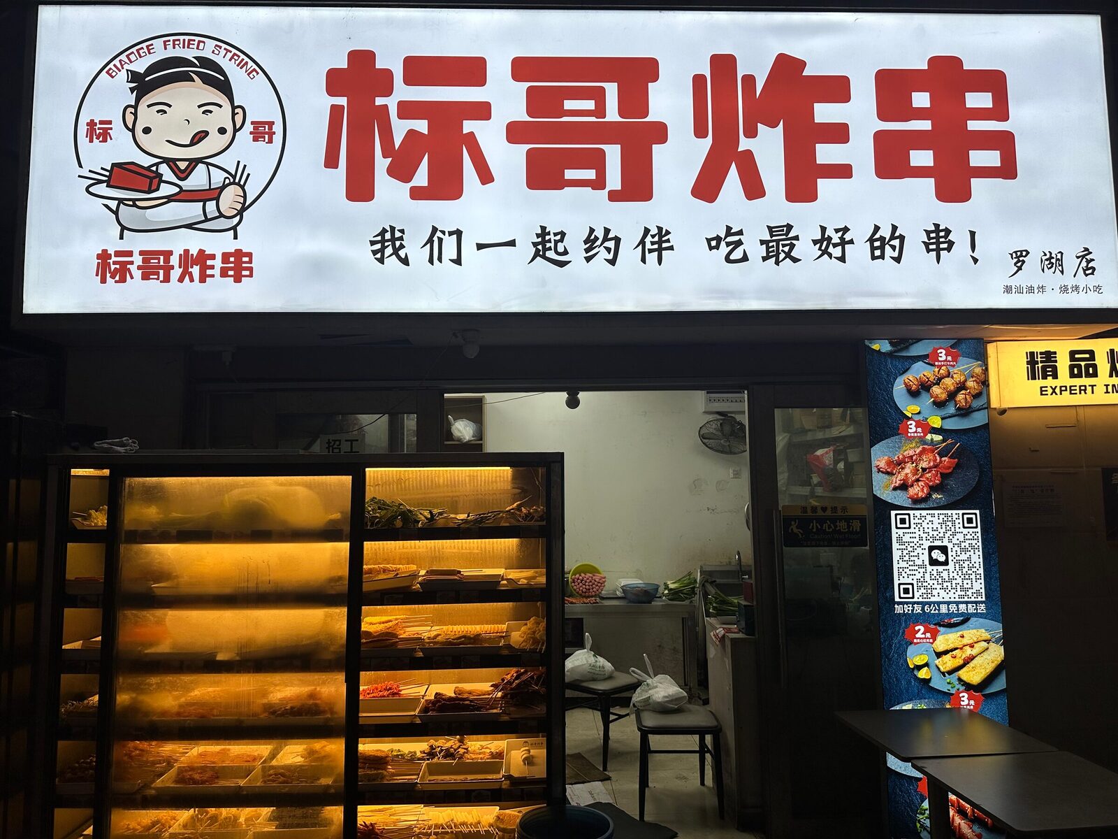Ett gatukök i stadsdelen Luohu i Shenzhen på kvällen. Under en upplyst skylt med röda kinesiska tecken står en glasmonter med belysta hyllor av råa grillspett, och till höger står en rollup med bilder på rätterna och QR-koder för beställning och betalning.