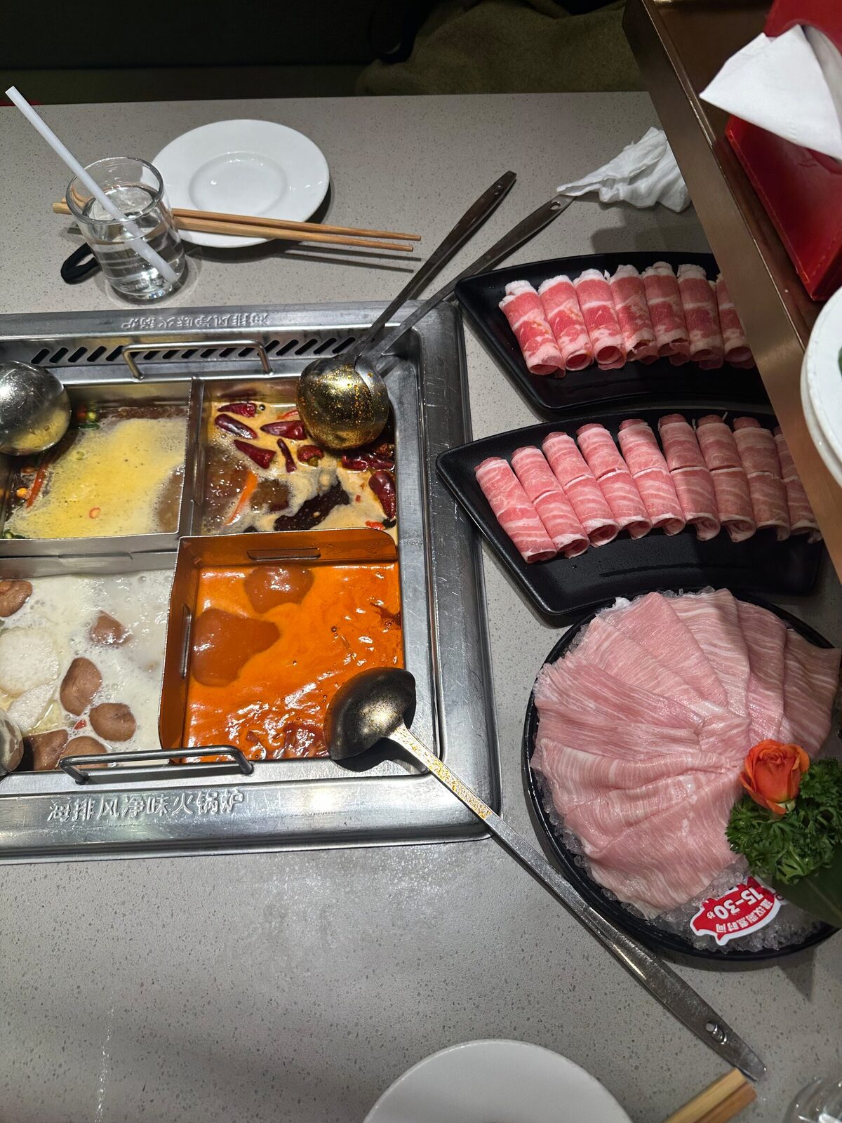 En serverad hotpot på ett restaurangbord med en fyrdelad gryta i bordets inbyggda häll, fylld med fyra olika buljonger, samt tallrikar med tunt skivat rått kött, slevar och ätpinnar bredvid.
