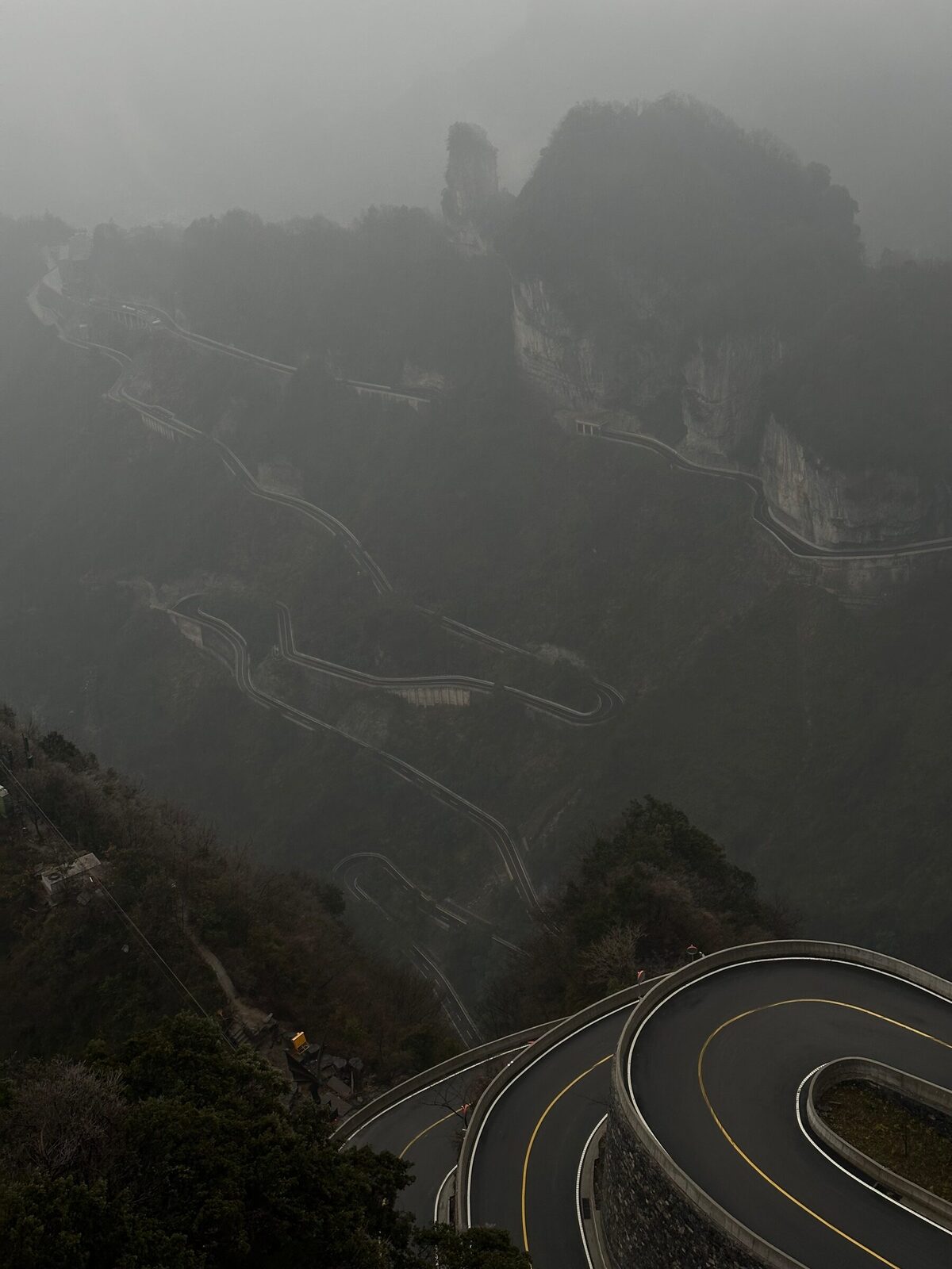 Bergsvägen upp mot Tianmen i Hunan sedd uppifrån i tät dimma, där asfaltsvägen slingrar sig i täta hårnålskurvor nedför bergssidan med gula mittlinjer och räcken längs kanten.