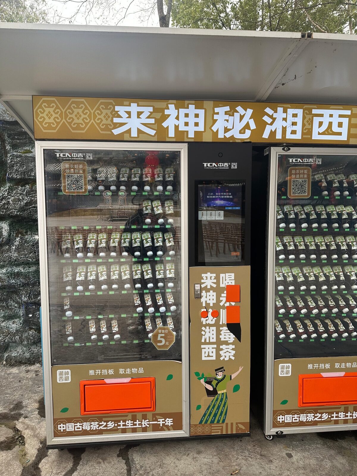 Närbild på två varuautomater i Xiangxi med produkthyllor fulla av flaskor med lokalt bärte. På vänstra dörren sitter en stor QR-kod för betalning, och mellan maskinerna sitter en smal videoskärm.