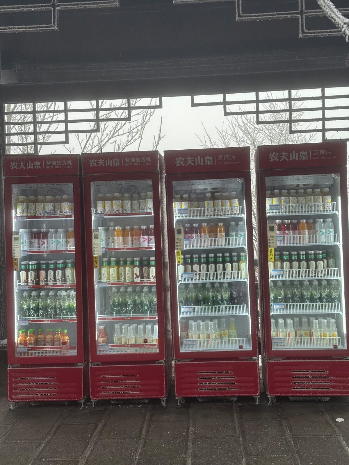 Närbild på fyra röda kylskåp med glasdörrar uppställda mot en frostig träfasad i Zhangjiajie. Hyllorna är fulla av flaskor med vatten, te och juice, och skyltarna längst upp anger att det rör sig om smarta varuautomater.