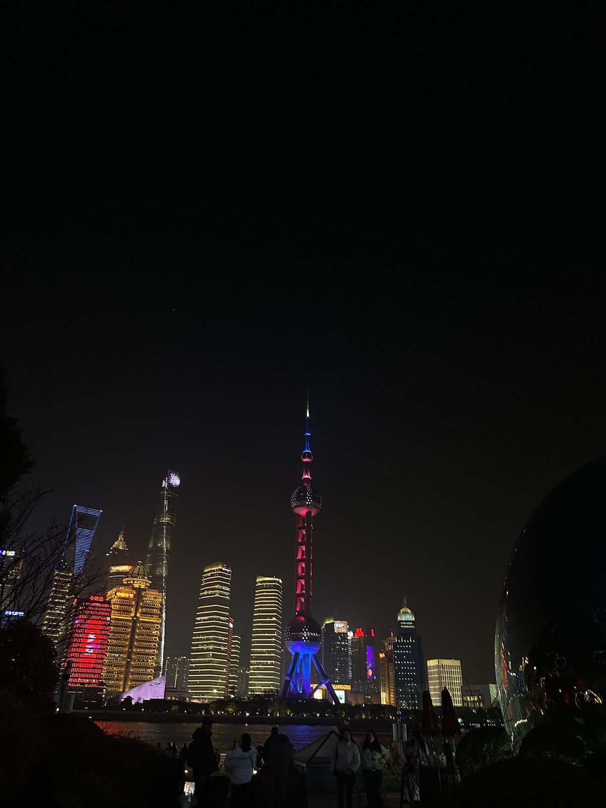 Skyskraporna i Pudong i Shanghai sedda från the Bund en kväll. Oriental Pearl Tower lyser i rosa och blått mitt i bilden, omgiven av upplysta höghus, och siluetter av åskådare syns längs kajen i förgrunden.