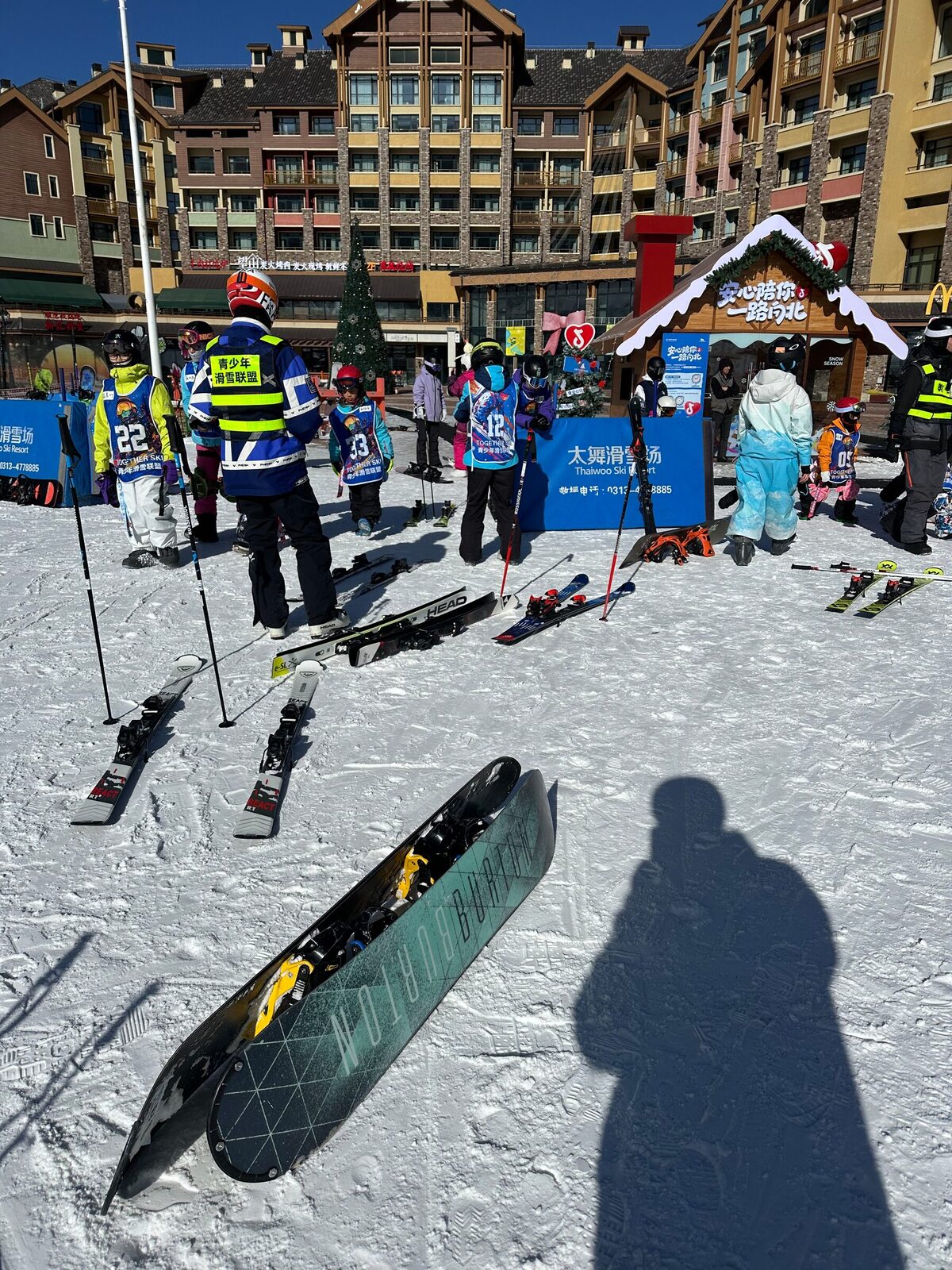 Basområdet på skidorten Thaiwoo i Hebei en solig vinterdag. Barn i numrerade västar samlas kring instruktörer i gula reflexvästar, skidor och stavar står nerstuckna i snön och alpinliknande hotellbyggnader reser sig i bakgrunden.