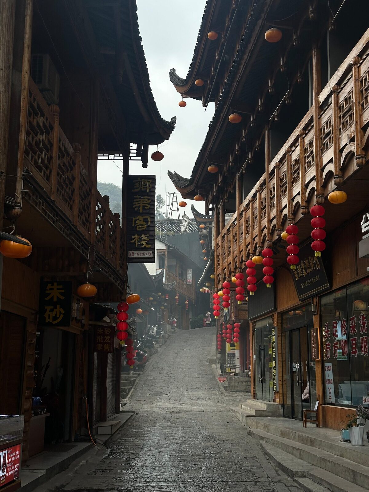 En smal stenlagd gränd i en gammal stadskärna i Xiangxi, kantad av traditionella trähus med snidade balkonger och utskjutande tak. Röda och orange lyktor hänger i rader längs fasaderna och butiksskyltar på kinesiska syns på båda sidor.