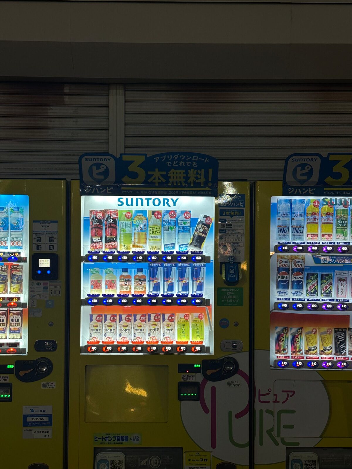 Närbild på tre gula dryckesautomater i rad framför en nerdragen butiksjalusi i Tokyo nattetid. Den mittersta bär Suntorys logotyp och har en kampanjskylt högst upp, och alla tre har tre upplysta hyllplan med flaskor och burkar bakom glaset.