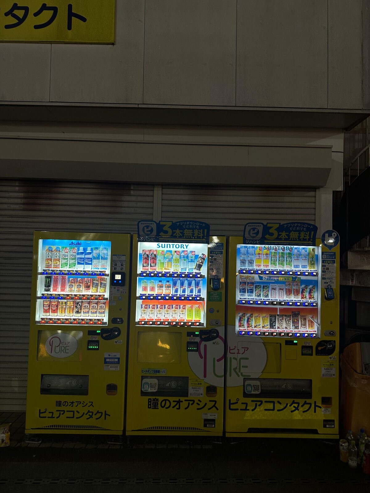 Tre gula dryckesautomater uppställda i rad framför en nerdragen butiksjalusi på en gata i Tokyo nattetid. Automaternas produkthyllor lyser upp trottoaren och visar rader av flaskor och burkar bakom glaset.