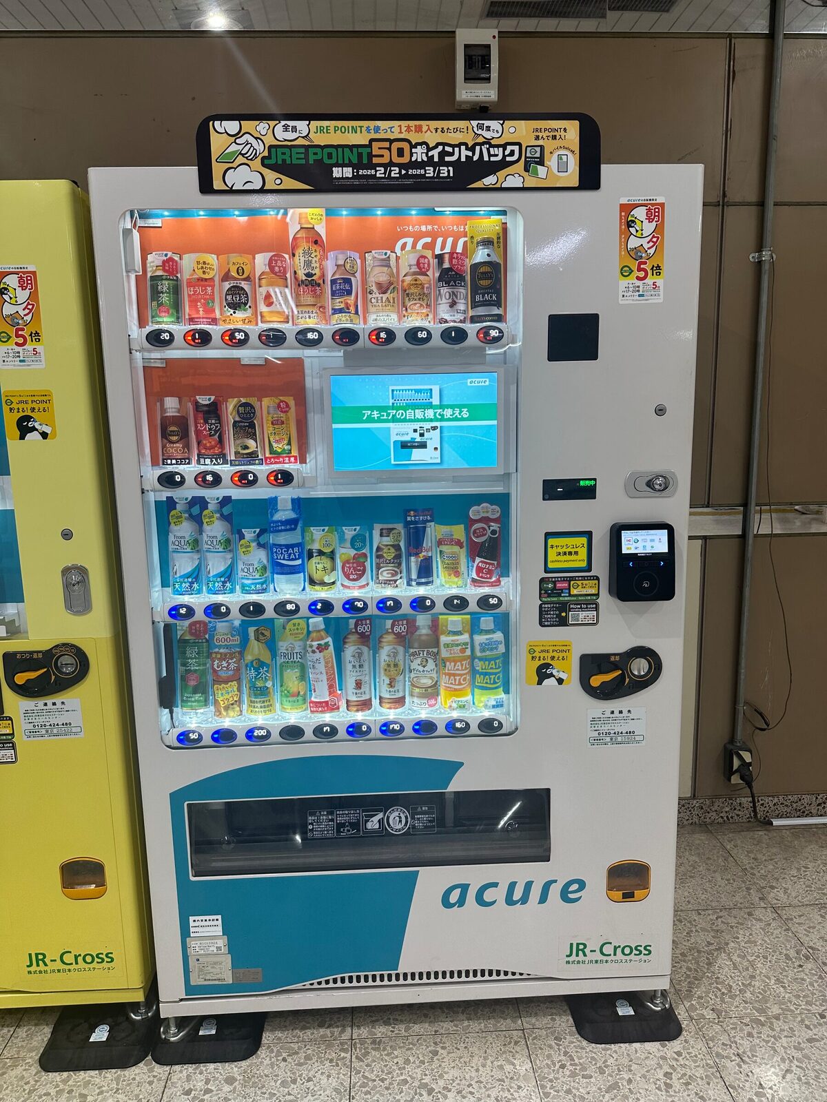 Dryckesautomat i ett köpcentrum med fyra hyllplan. Det översta hyllplanet är belyst i varm orange ton och innehåller burkar med kaffe och te, medan hyllplanen under är belysta i kallt blått ljus och innehåller vatten och läsk.