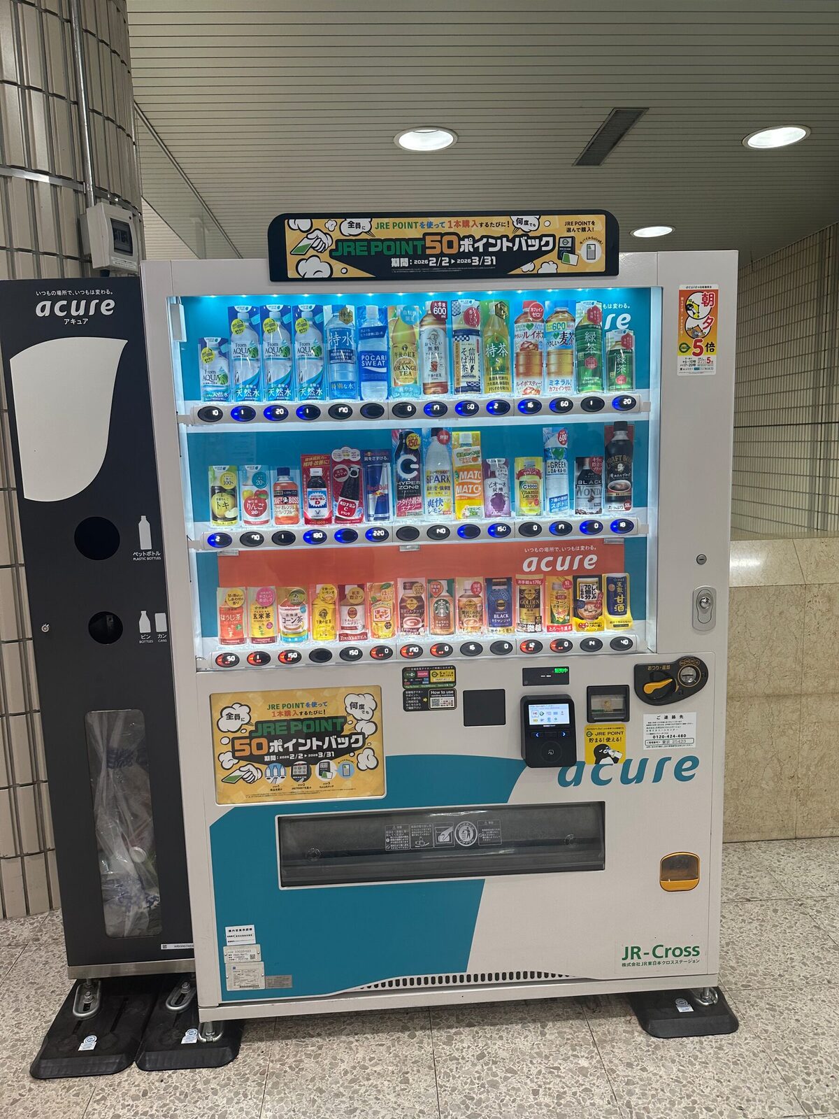 Vit och turkos dryckesautomat i en tunnelbanestation i Tokyo med tre upplysta hyllplan fulla av flaskor och burkar. Ovanpå maskinen sitter en reklamskylt för ett poängprogram, och till vänster står en matchande återvinningsbehållare för flaskor.