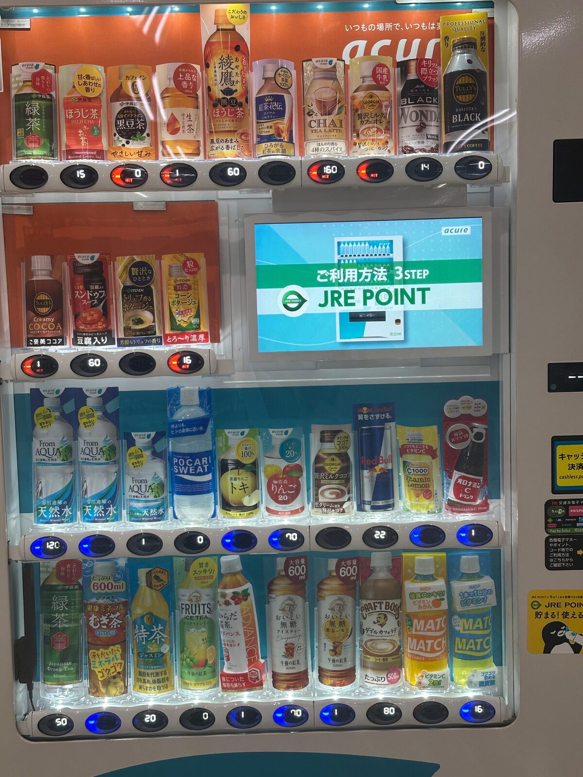 Närbild på automatens hyllplan där skillnaden syns tydligt: överst rader med varma burkar te och kaffe mot orange bakgrund med röda knappar, därunder en informationsskärm, och nederst kalla flaskor med vatten, sportdryck och juice med blå knappar.