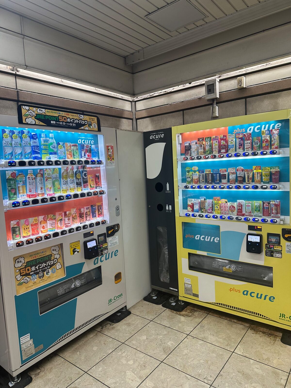 Två dryckesautomater från samma operatör står i en innerhörna i en japansk byggnad, den ena ljusblå och den andra gul, båda med upplysta produkthyllor och betalterminaler.