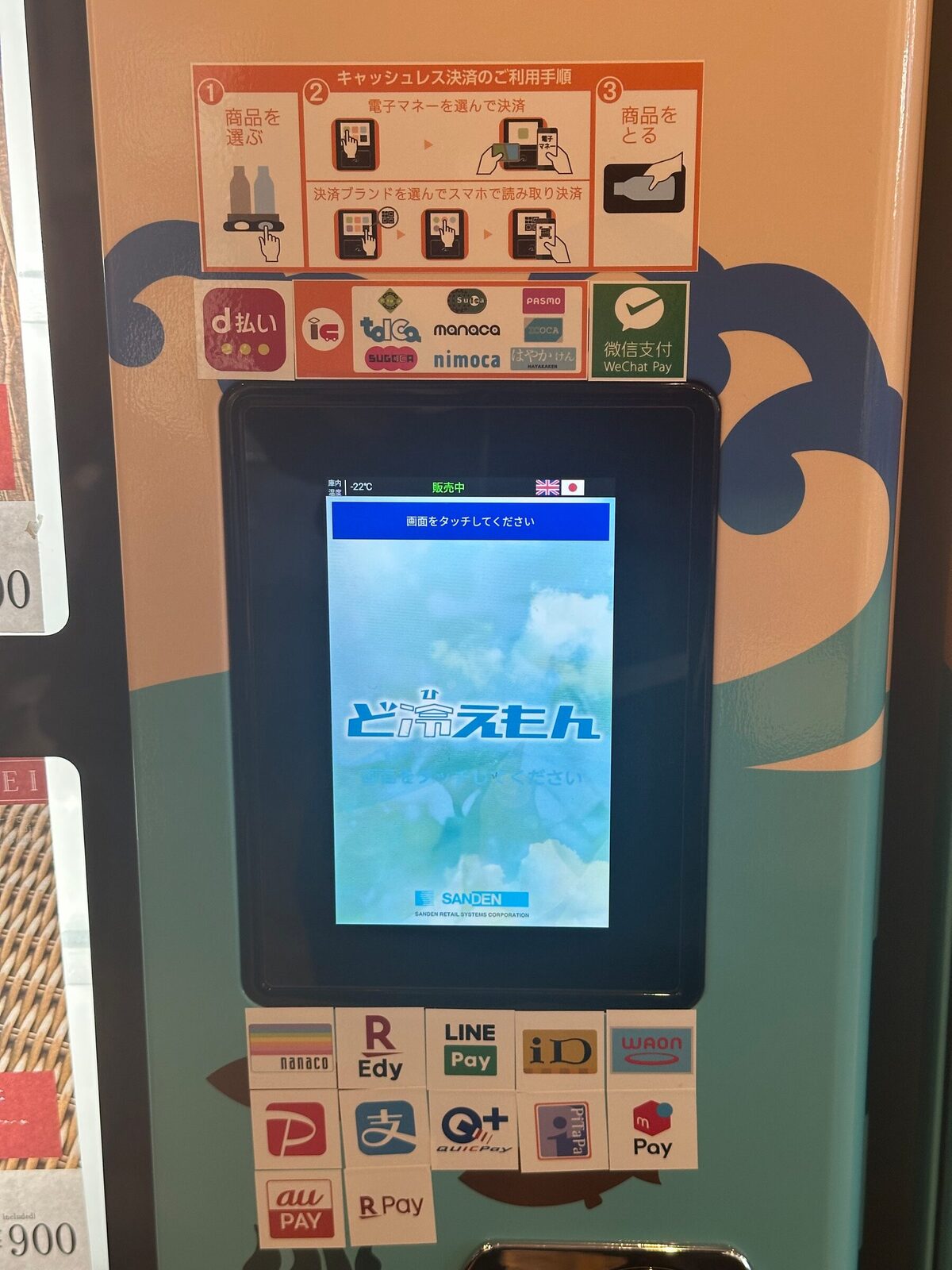 Närbild på betalningsdelen av en japansk dryckesautomat. Överst sitter en instruktionsskylt i tre steg med bilder, därunder en pekskärm med språkval, och runt omkring ett drygt tjugotal dekaler med logotyper för olika elektroniska plånböcker och betalappar.