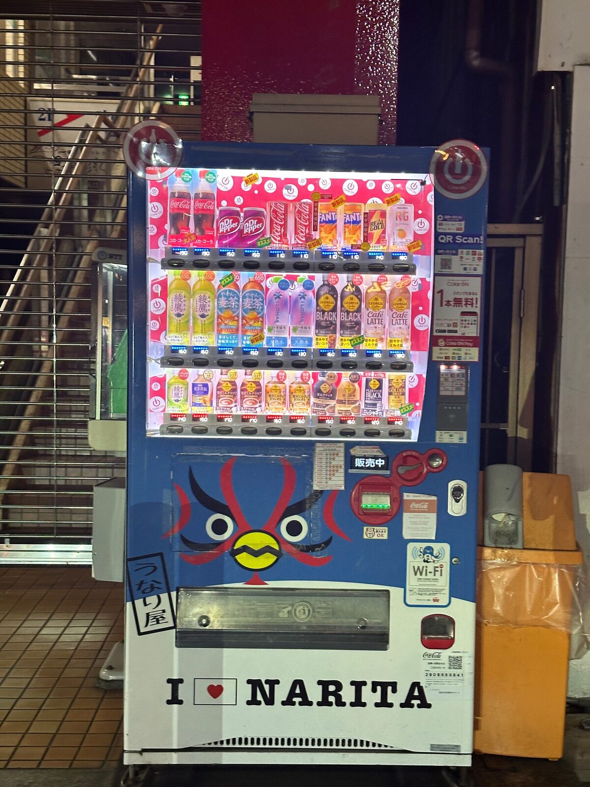 En dryckesautomat i Narita dekorerad med ett målat rött demonansikte och texten I love Narita, med rader av flaskor och burkar bakom glaset, placerad under en yttertrappa.