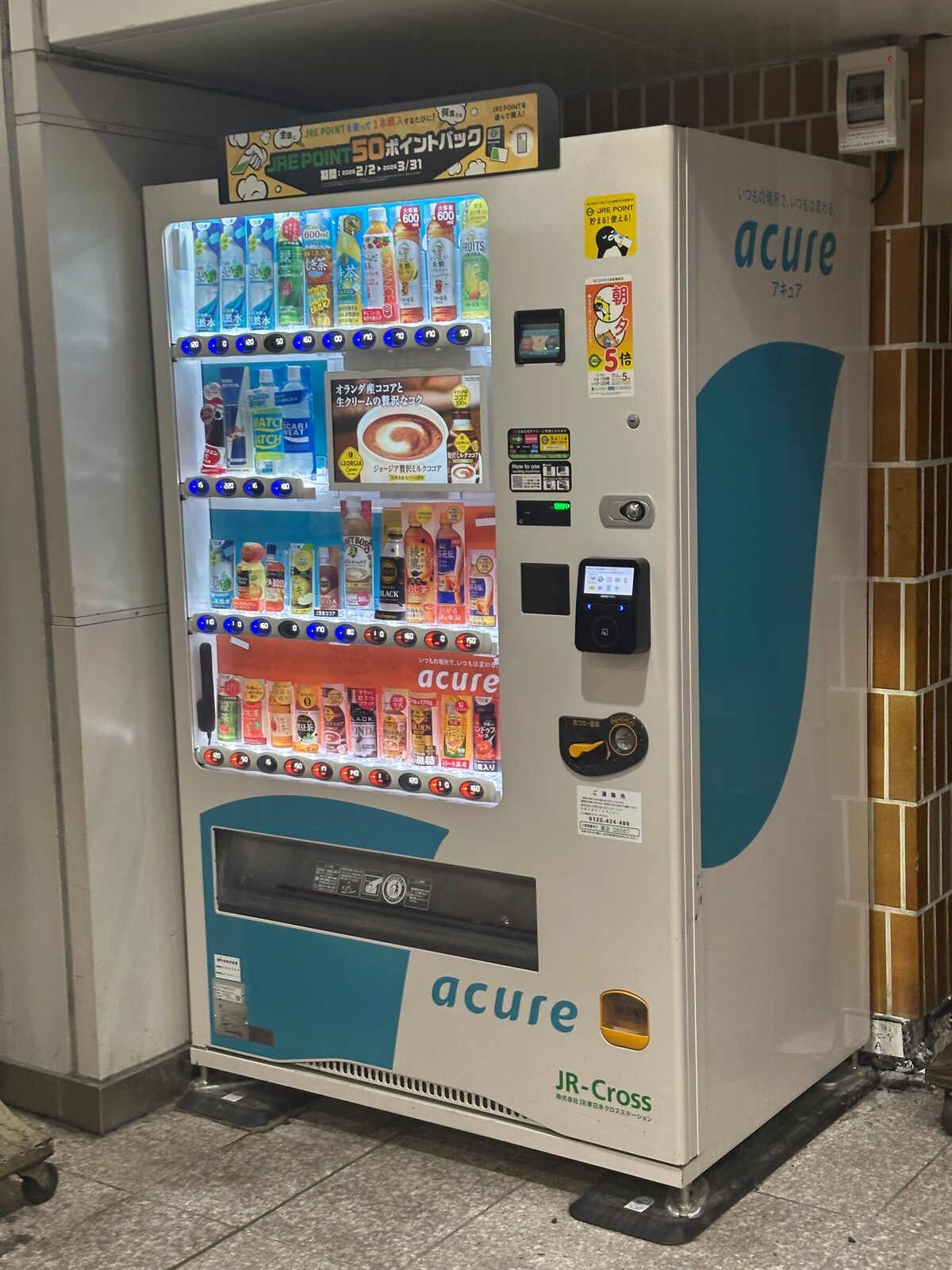 Vit och turkos dryckesautomat i en tunnelbanestation i Tokyo, med en kampanjskylt för järnvägens poängprogram högst upp, upplysta hyllor med flaskor, en liten reklamskärm i mitten och en betalterminal till höger.