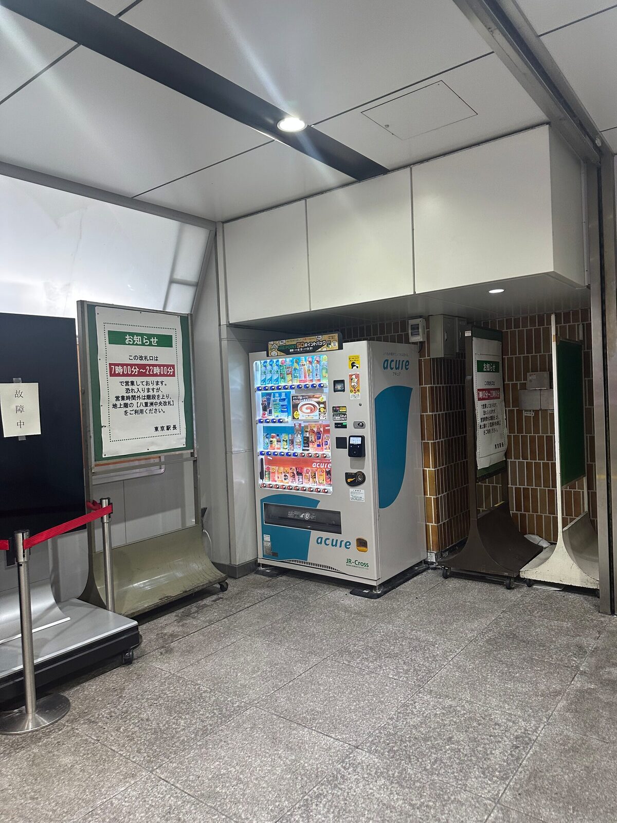En dryckesautomat placerad i en nisch intill en mindre stationsutgång i Tokyo. Bredvid hänger en anslagstavla med öppettider för utgången, och avspärrningsstolpar med rött band står på stengolvet framför.
