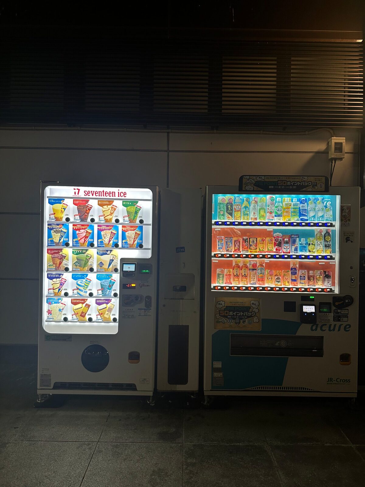 En glassautomat och en dryckesautomat står bredvid varandra mot en mörk betongvägg med ventilationsgaller ovanför. Båda maskinernas produkthyllor lyser upp det omgivande mörkret.