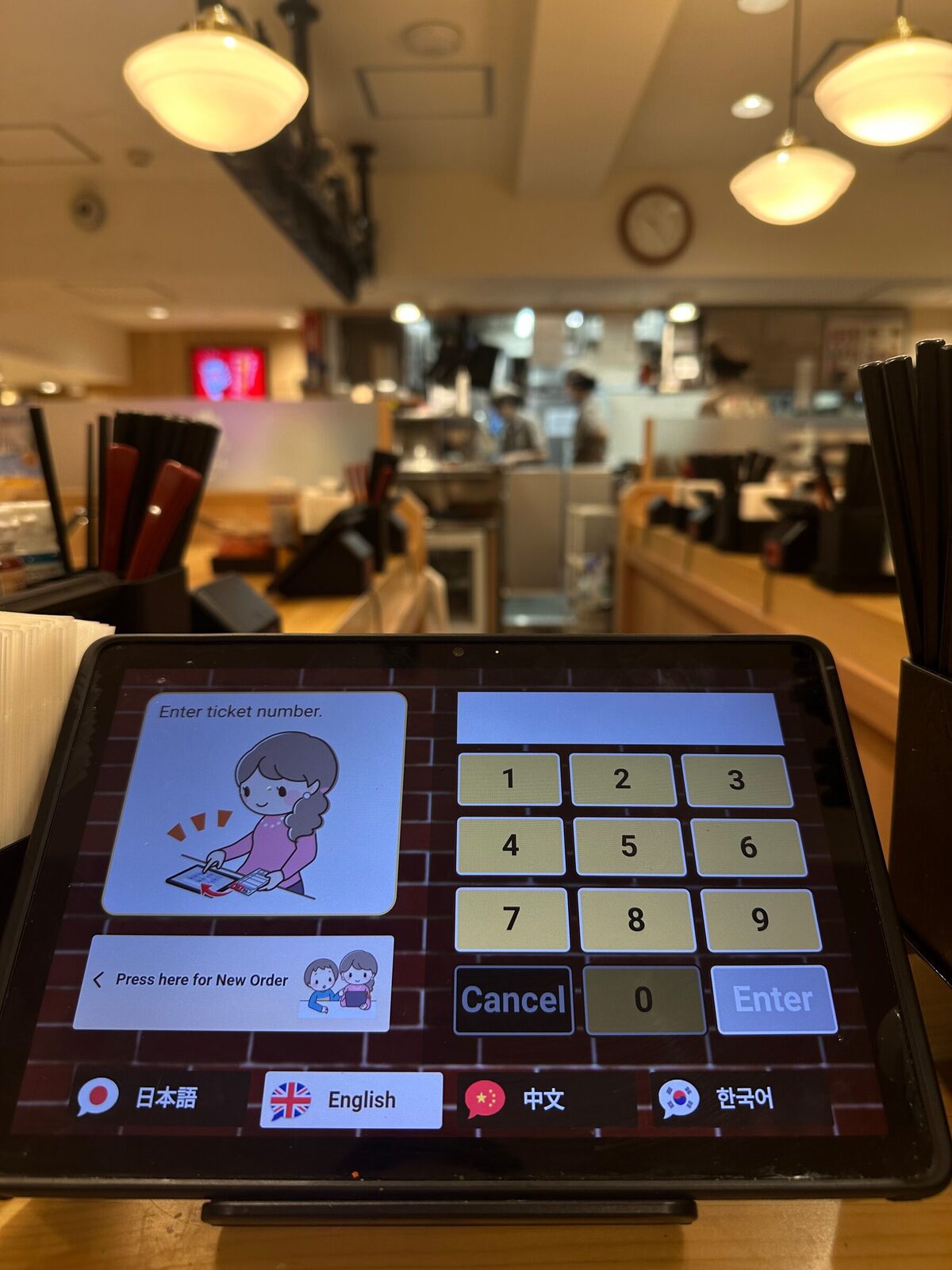 En pekskärm på en disk i en japansk restaurang visar ett numeriskt tangentbord med texten Enter ticket number och knappar för att växla språk mellan japanska, engelska, kinesiska och koreanska. I bakgrunden syns det öppna köket och hängande taklampor.