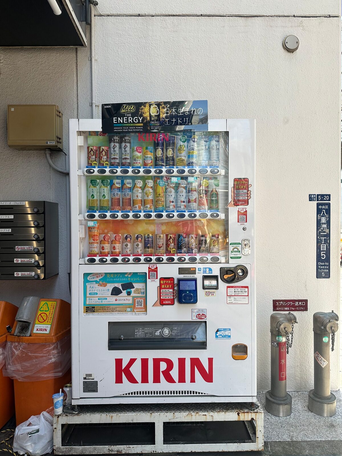 En vit dryckesautomat märkt Kirin står mot en putsad husvägg i Tokyo, med rader av flaskor och burkar bakom glaset. Till vänster sitter en rad postfack på väggen och till höger står två brandposter.