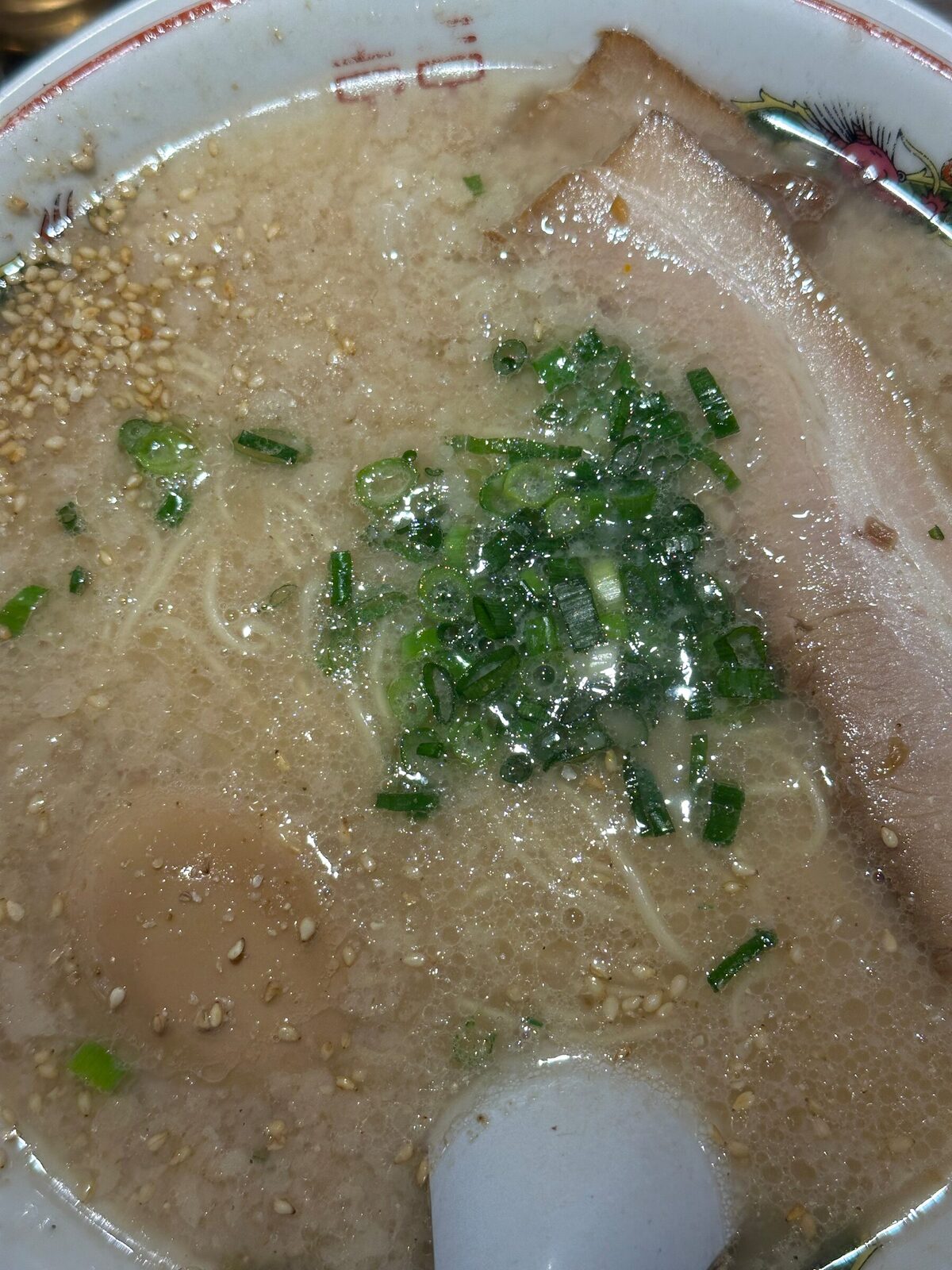 Närbild uppifrån på en skål tonkotsu-ramen med grumlig ljus buljong, tunna nudlar, en skiva fläskkarré, hackad salladslök, sesamfrön och ett halvt kokt ägg.