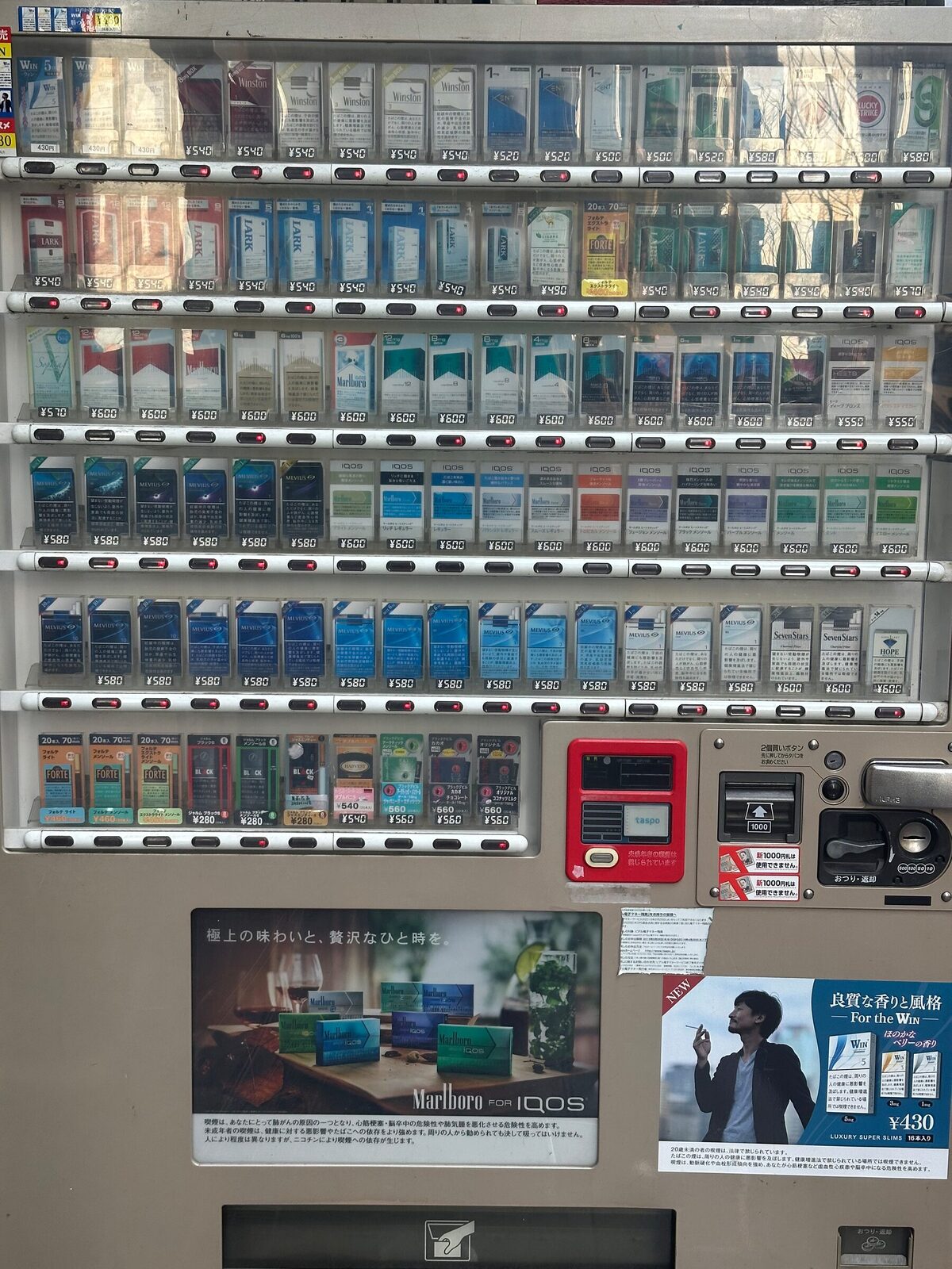 Närbild på fronten av en japansk tobaksautomat i Tokyo. Bakom glaset står cigarettpaket i sex rader med prislappar i yen, och till höger sitter en röd taspo-läsare för åldersverifiering intill sedelinkast och myntinkast.