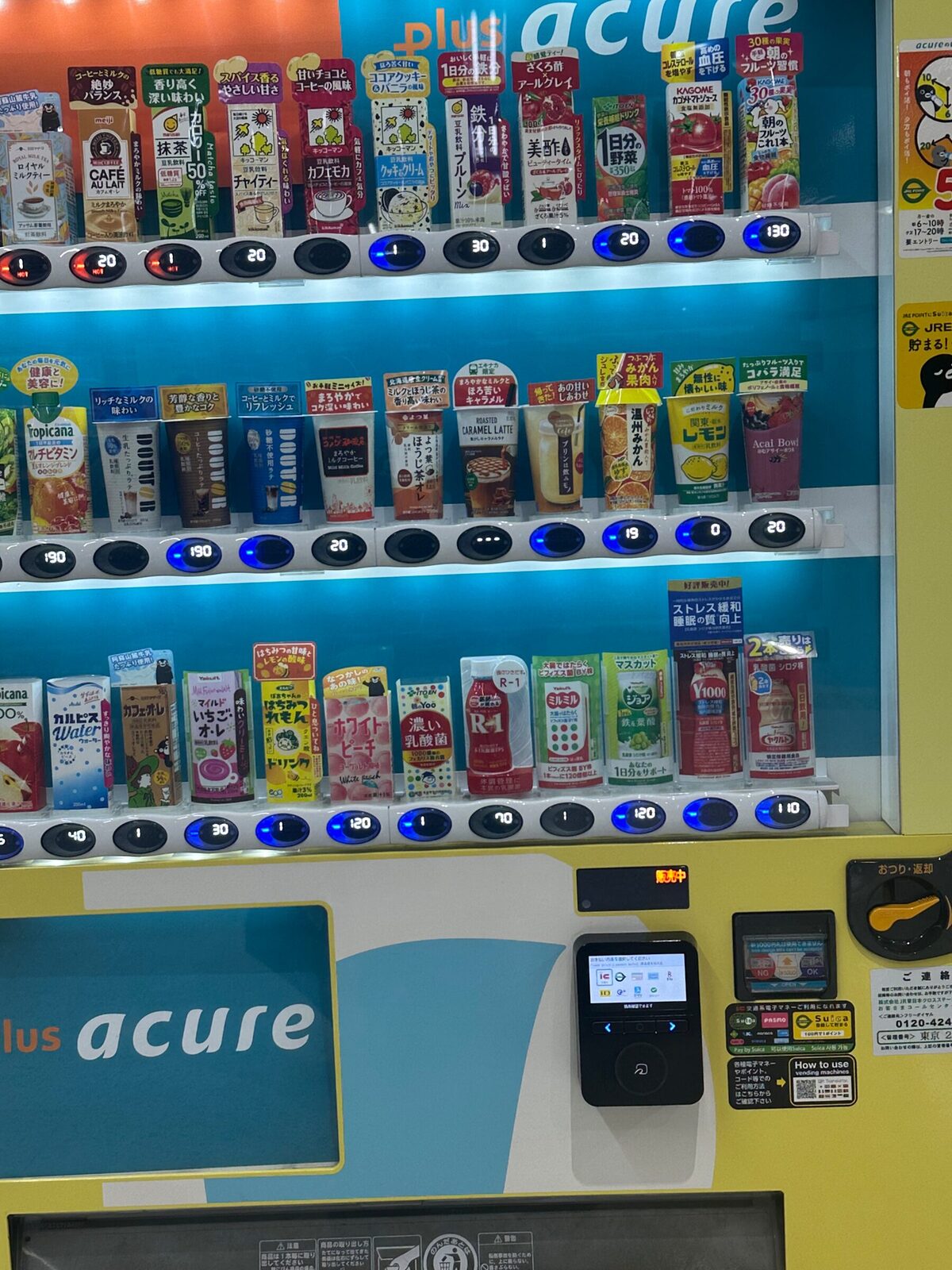 Närbild på den gula automatens hyllplan, som nästan uteslutande innehåller pappförpackningar: mjölkte, grönt te, grönsaksjuice, kaffe och yoghurtdrycker, med prisknappar under varje rad och en betalterminal längst ner till höger.
