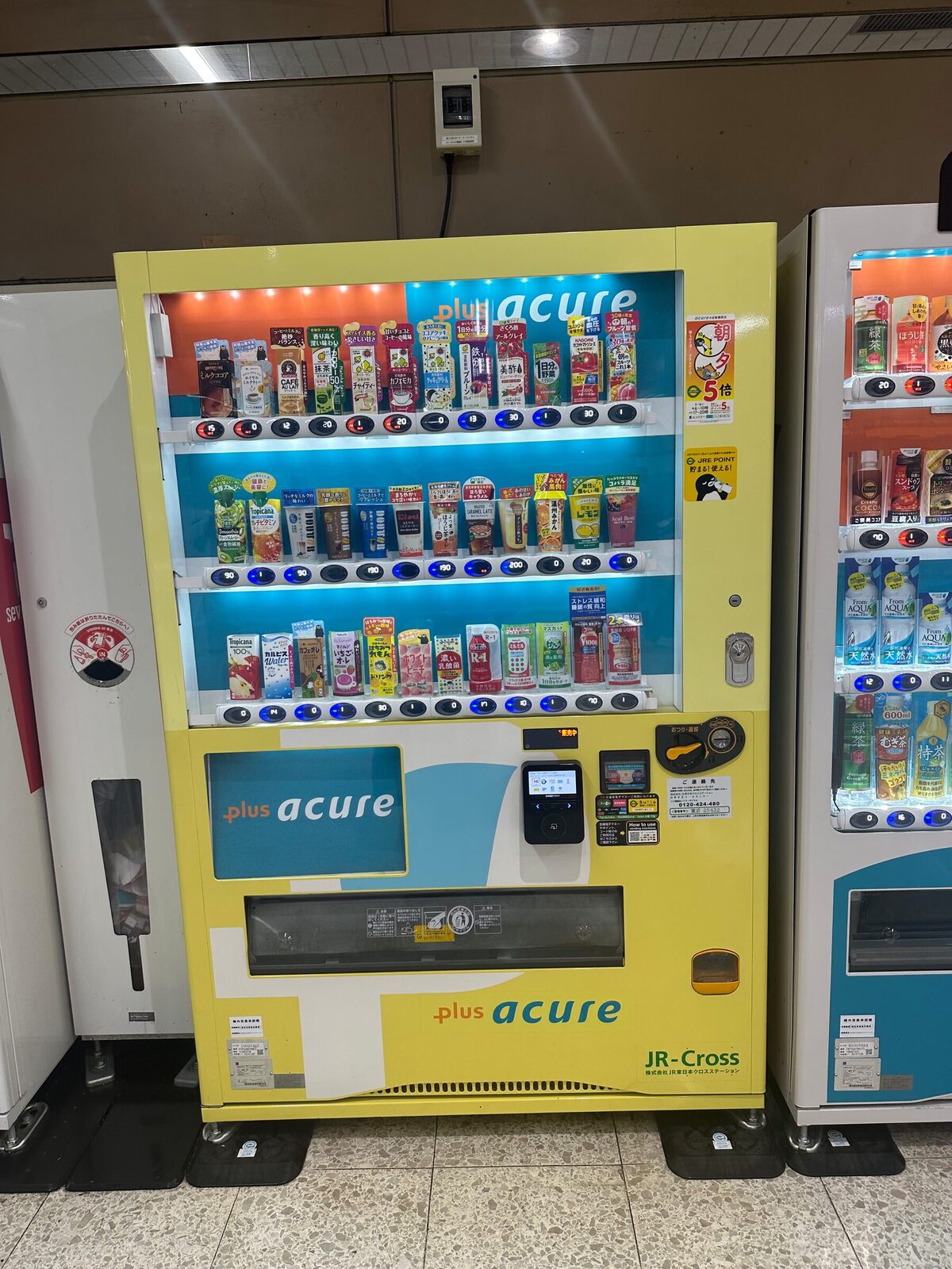 Gul dryckesautomat i en tågstation i Tokyo med tre hyllplan som mestadels innehåller pappförpackningar och hälsodrycker istället för läsk. Bredvid står ytterligare en automat med ett annat sortiment.