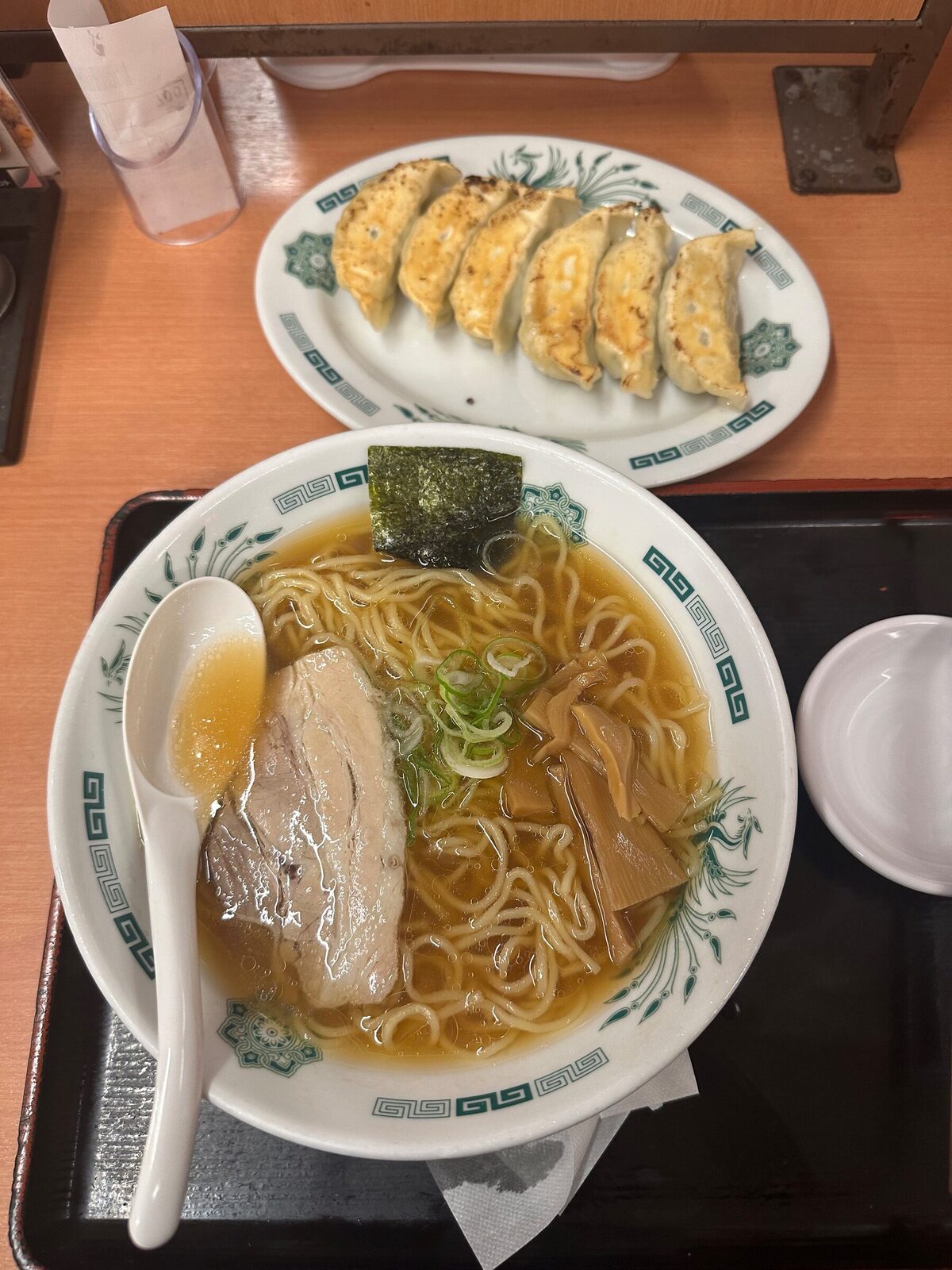 En bricka med en skål shoyu-ramen med klar brun buljong, nudlar, en skiva fläskkarré, bambuskott, salladslök och ett ark nori, samt ett fat med sex stekta gyoza bredvid.