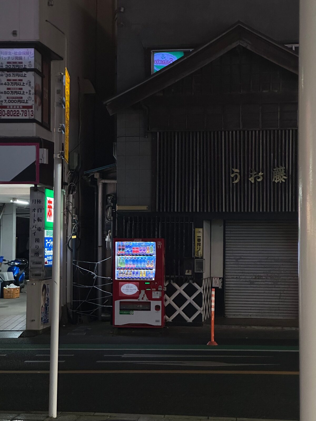 En ensam röd och vit dryckesautomat lyser i mörkret intill ett traditionellt japanskt trähus med nerdragen plåtjalusi på en tyst gata nattetid. Automaten är den enda ljuskällan på gatuplanet.