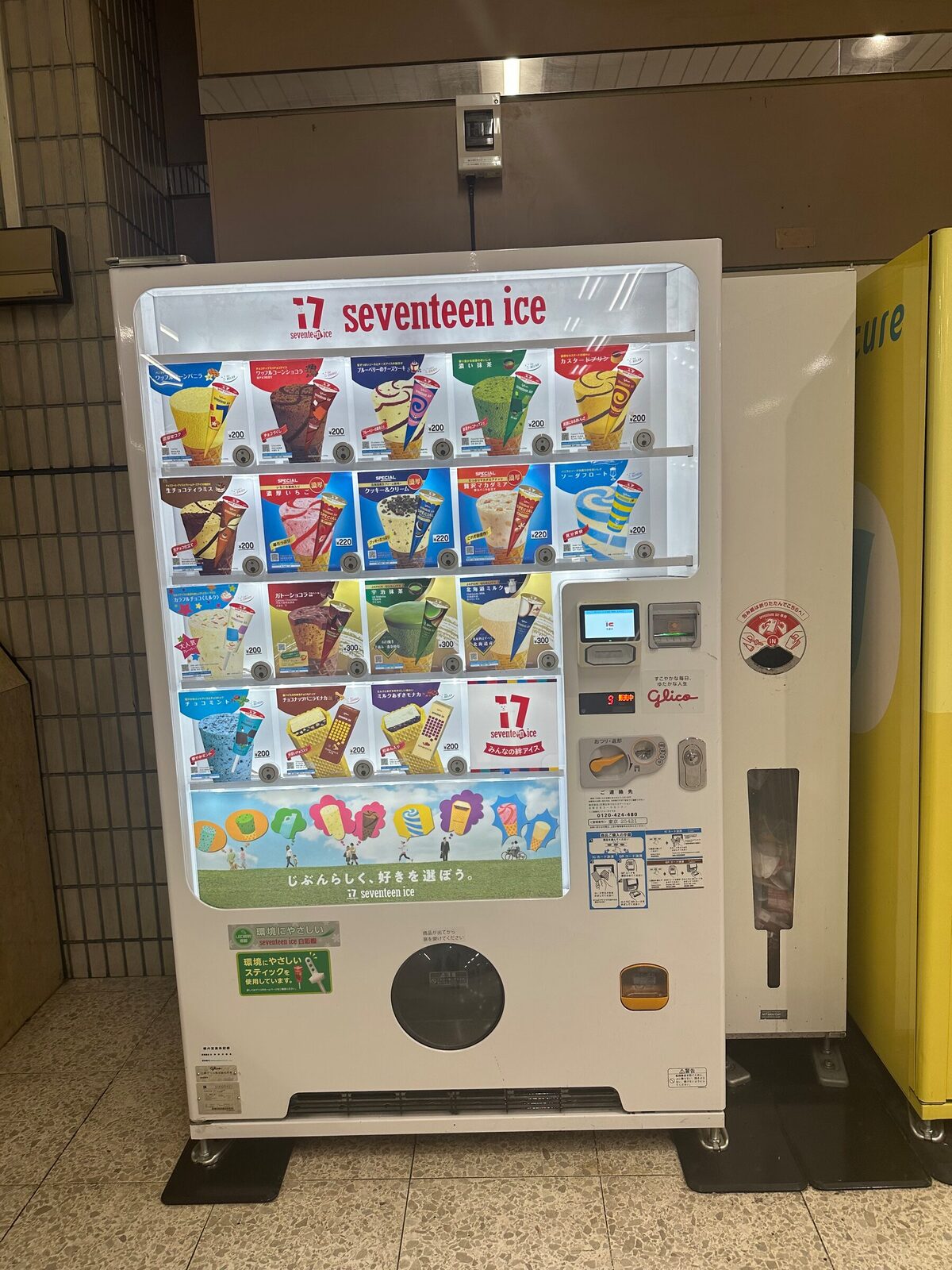 Vit glassautomat i en station i Tokyo med fyra hyllplan som visar ett tjugotal olika glassorter i strut- och stickform, alla märkta med samma pris. Nedtill sitter en rund utlämningslucka och till höger en betalterminal.