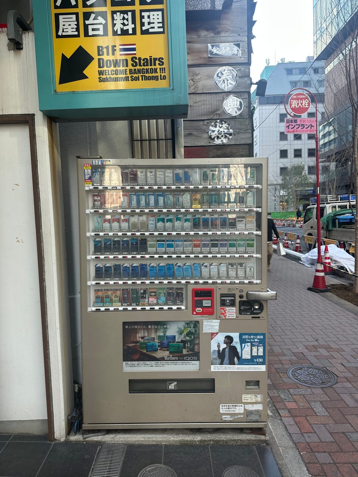 Tobaksautomat som står fritt på trottoaren längs en gata i Tokyo, inklämd mellan en husvägg och gångbanan. Ingen personal finns i närheten och fotgängare passerar förbi på trottoaren bakom.