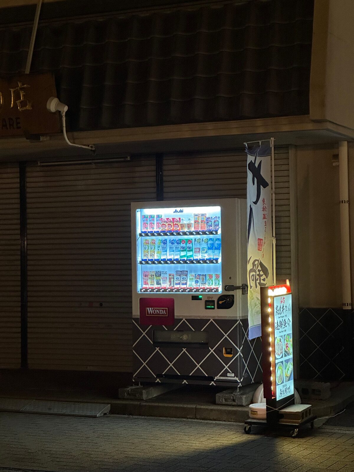 En upplyst varuautomat står ensam framför en stängd butik med nerdragen jalusi på en gata i Tokyo på kvällen. Bredvid automaten står en lysande reklamskylt, och gatan är tom.