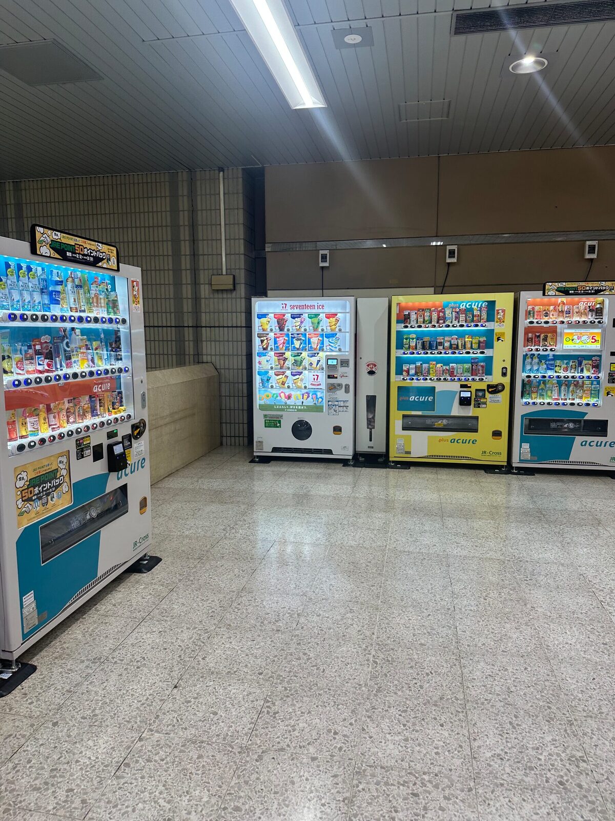 Tre varuautomater står uppradade längs väggen i en japansk tunnelbanestation, med upplysta produkthyllor mot ett tomt terrazzogolv under lysrörsbelysning.