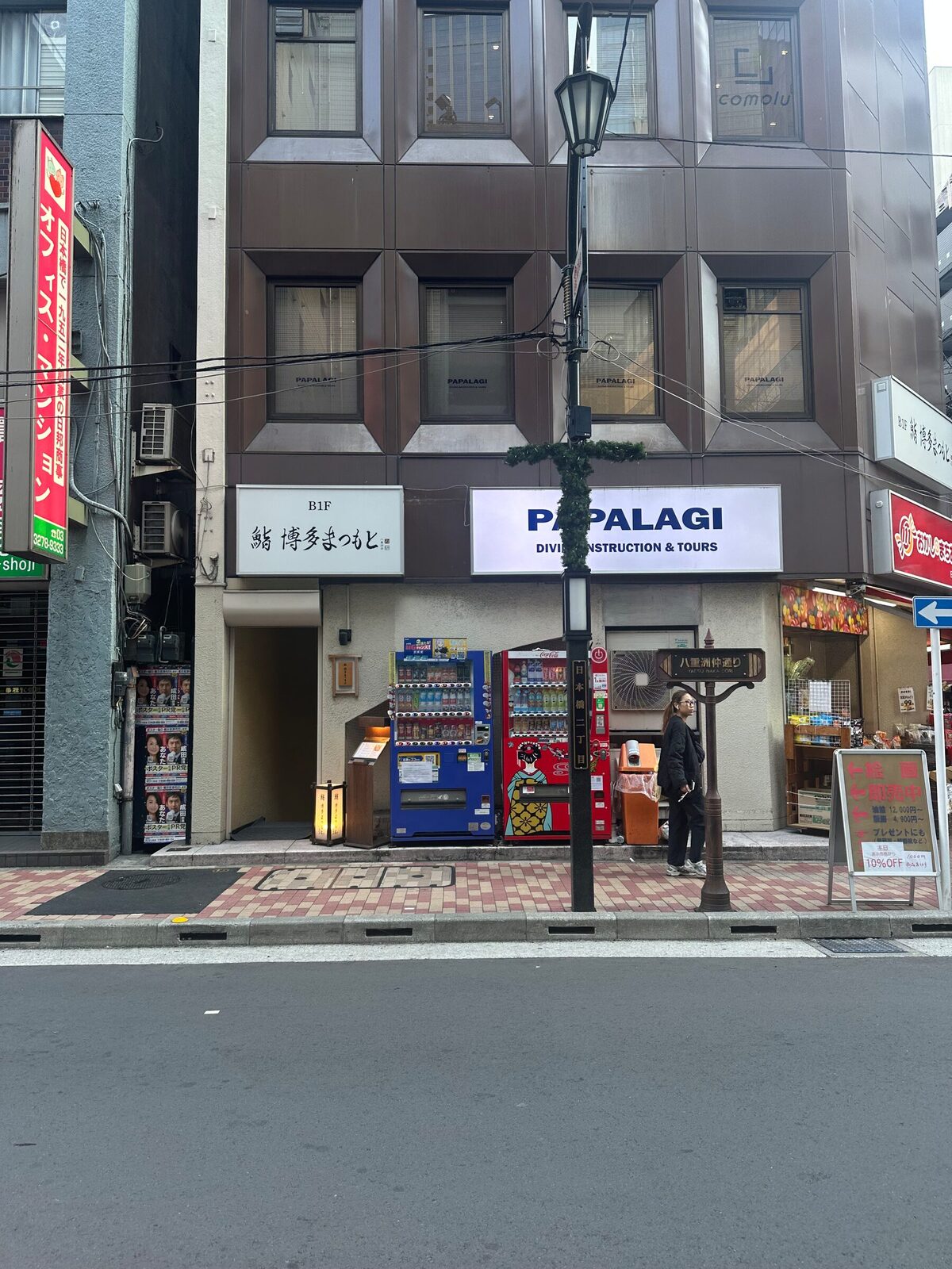 En gata i Tokyo där flera varuautomater står intill varandra i bottenvåningen på en byggnad, med en fotgängare på trottoaren och butiksskyltar ovanför.