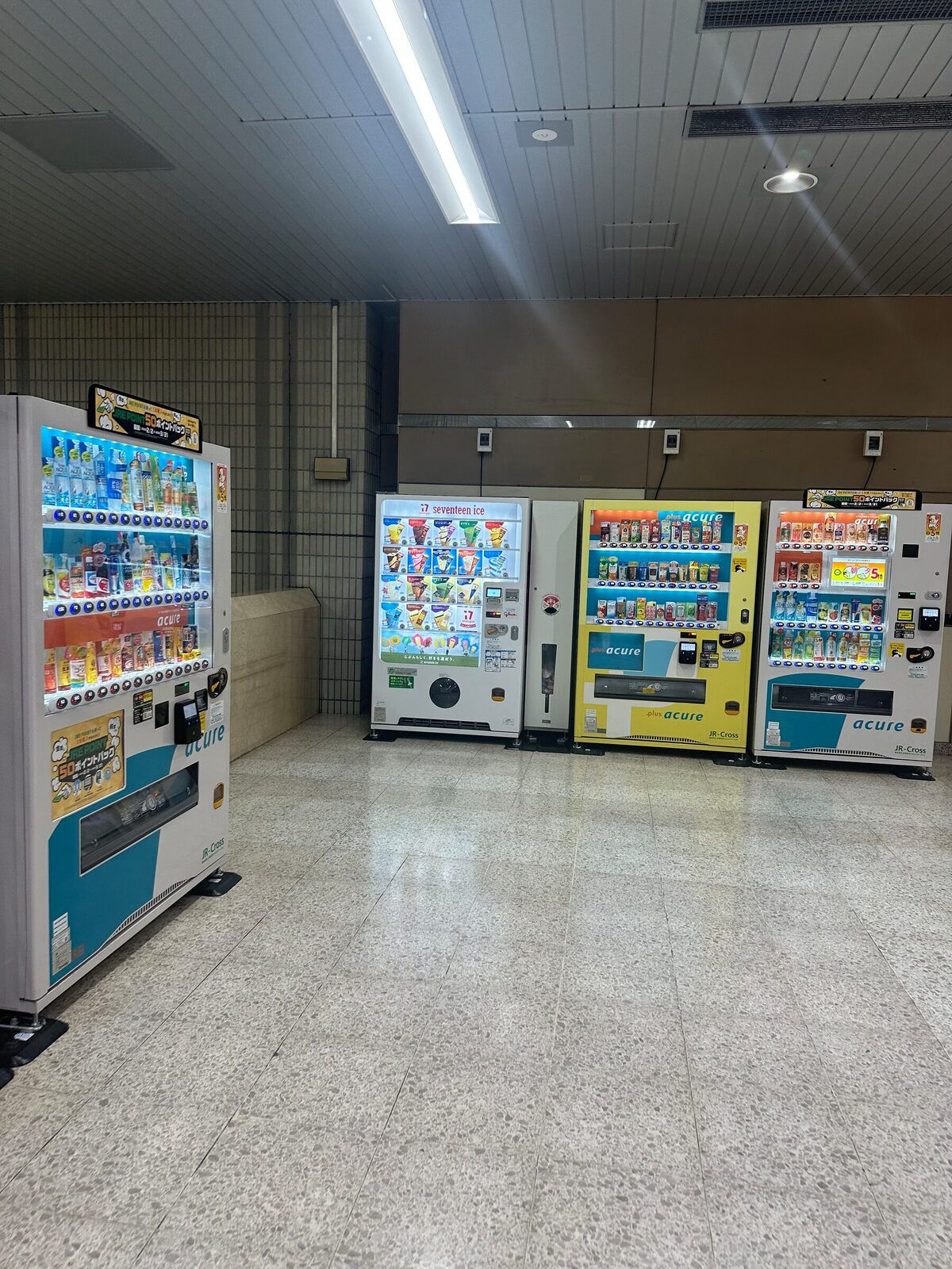 Fem vendingmaskiner uppradade längs väggen i en tunnelbanestation i Tokyo. Längst till vänster en hög dryckesautomat, därefter en glassautomat och tre dryckesautomater med upplysta produkthyllor, alla på ett tomt stengolv under lysrörsbelysning.